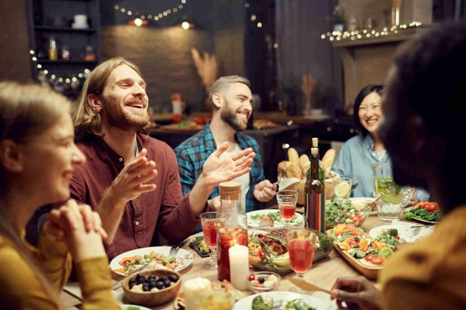 A group of friends gathered at a dinner table out at a restaurant enjoy food and drinks while talking and laughing. 