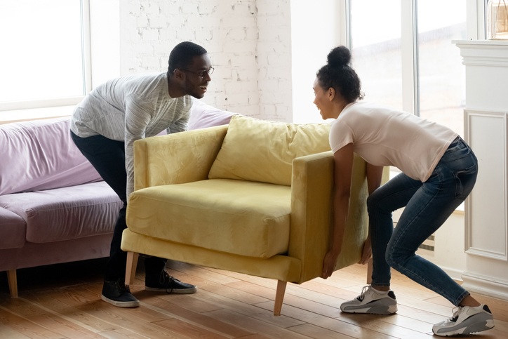 Two people lift a plush chair to move it in a small room