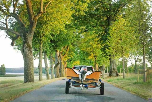 A white vehicle pulls a yellow boat down a road surrounded by trees