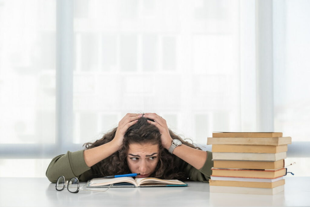 Woman with her hands on her head and a stressed facial expression sitting at a table with a stack of books.