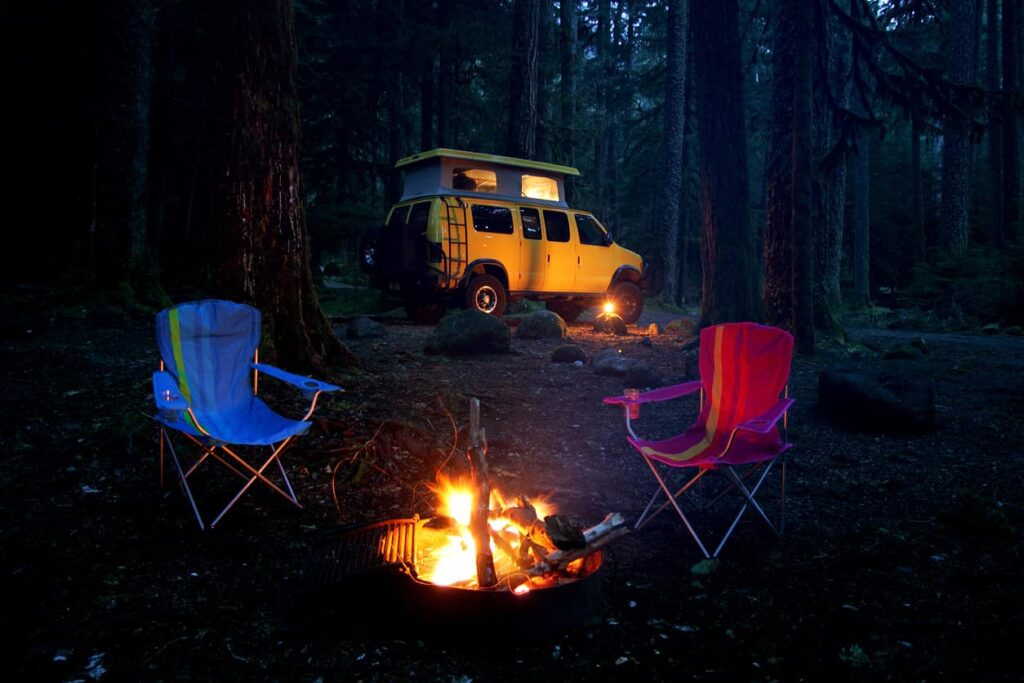 Campervan parked behind a forest-surrounded campfire and two lawn chairs at dusk.