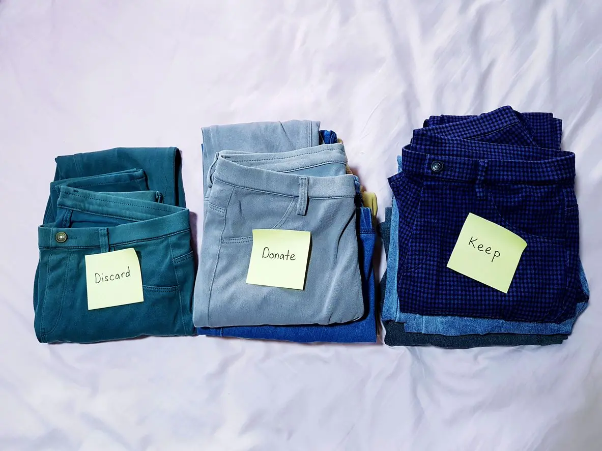 Three stacks of folded pants sit on a bed, with sticky notes reading “discard,” “donate,” and “keep.”