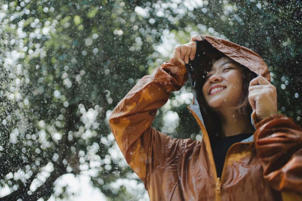 A young woman smiles at the rain while holding her hood. 