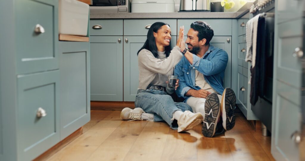 A couple sitting down on the kitchen floor, high-fiving. 